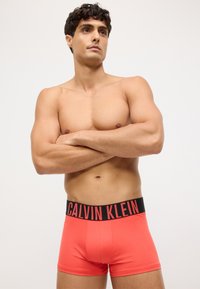 Crvene Calvin Klein bokserice s crnim pojasom na kojem se u podebljanim crvenim slovima nalazi ime branda. Glatki, rastezljivi materijal.
