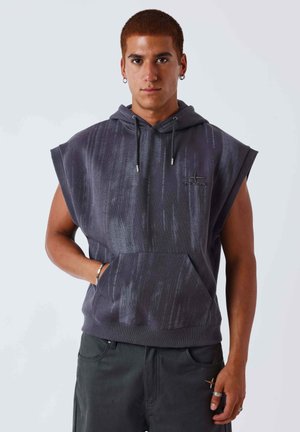Jeune homme aux cheveux courts portant un sweat à capuche sans manches gris foncé avec poche avant et pantalon foncé, debout devant un fond clair uni.