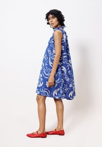 Blaues Blumenbekleidungsstück mit lockerer Passform, aus leichtem Stoff gefertigt. Es verfügt über einen Neckholder-Ausschnitt und einen fließenden Rock. Kombiniert mit roten flachen Schuhen.