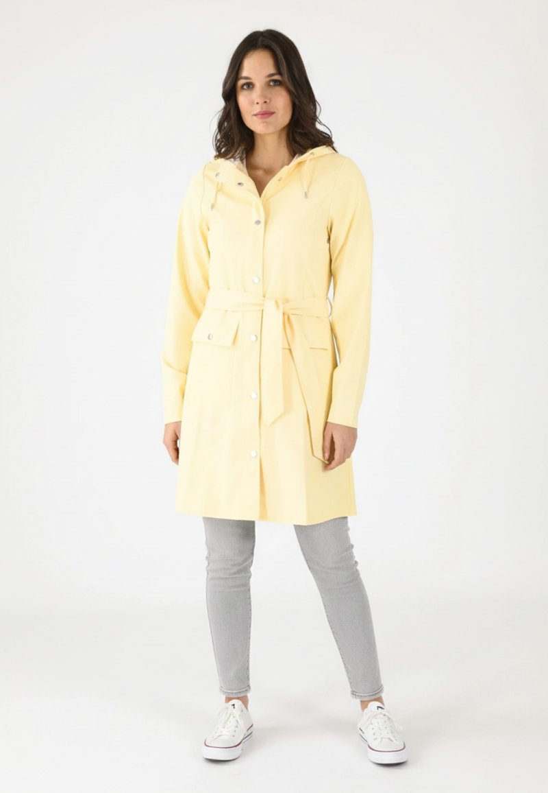 Imperméable jaune à boutons avec une capuche, ceinturé à la taille. Présente des poches avant et une texture lisse. Associé à un jean gris et des baskets blanches.