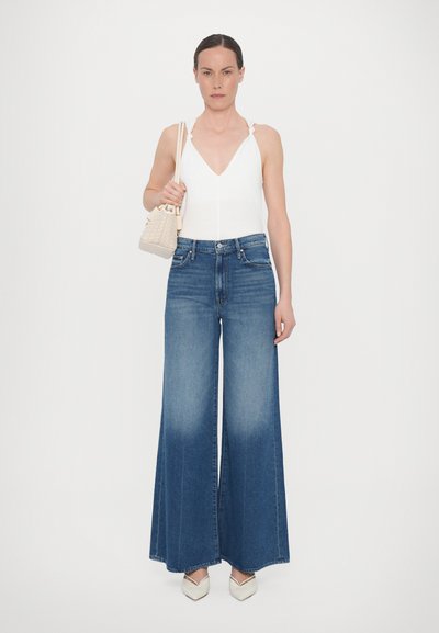 Wide-leg blue denim jeans with a light fade, paired with a white halter top and a small beige quilted handbag. Features pointed white shoes.
