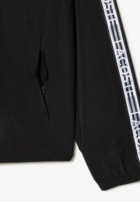 Svart jacka med en dragkedjeficka och elastiska ärmslut. Har vit "LACOSTE" logotyp vertikalt längs ärmen. Slät, lättviktsmaterial.