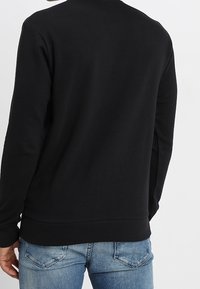 Svart sweatshirt med rund hals och ribbade ärmslut. Materialet verkar jämnt och det är kombinerat med ljusblå jeans, vilket visar en avslappnad stil.