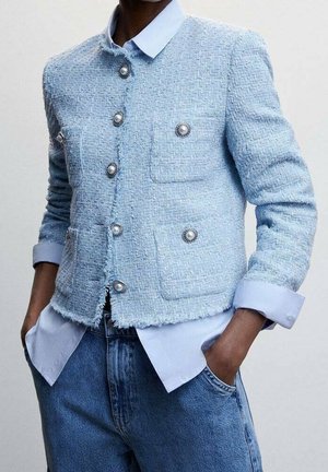 Light blue textured jacket with pearl and silver buttons worn over a pale blue shirt, paired with high-waisted blue jeans and hands in pockets.