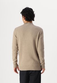 Mann mit lockigem, dunklem Haar, der einen beigen, strukturierten Strickpullover und schwarze Hosen trägt und vor einem schlichten weißen Hintergrund steht.