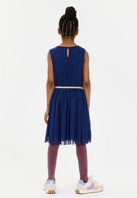 Abito blu navy con gonna svasata, accentuato da una cintura dorata e dettaglio a fessura sulla schiena. Indossato con leggings rosa brillanti e sneakers bianche.