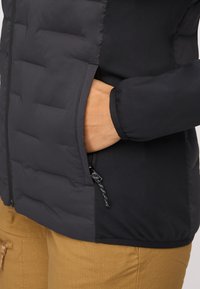 Svart isolerad jacka med quiltat mönster, utrustad med en ficka med dragkedja och släta tygdetaljer längs ärmarna; tillsammans med bruna byxor.