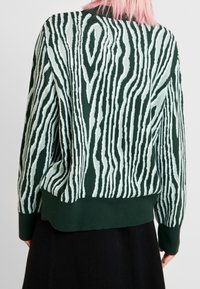 Grüner Pullover mit einem strukturierten Holzmaserungsmuster in Hellgrün, langen Ärmeln und einem gerippten Saum, getragen über einem schwarzen Rock.