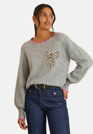 Grauer Strickpullover mit einem gewellten rosa Ausschnitt und einem Leopardenmuster-Schleifendetail. Kombiniert mit hochgeschnittenen dunkelblauen Jeans.