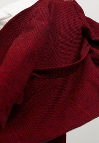 Punainen ja musta teksturoitu villatakki asetettuna valkoiselle pinnalle, näyttäen läheltä kankaan yksityiskohtia ja etutaskua.