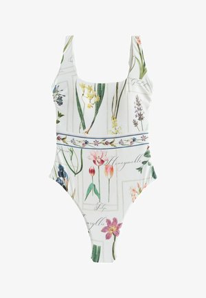 Femeie cu păr lung și blond, purtând un costum de baie alb cu flori, stând sprijinită de o suprafață de piatră texturată, uitându-se spre dreapta.