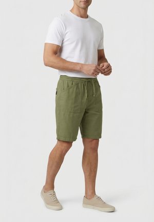 Uomo che indossa una maglietta bianca a maniche corte, pantaloncini verde oliva con coulisse e tasche frontali, e scarpe da ginnastica beige, in piedi davanti a uno sfondo neutro.