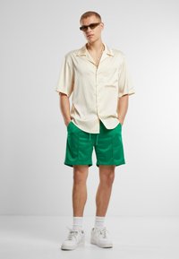 Kortærmet lyscreme skjorte med lomme, parret med grønne træningsshorts. Fodtøj er hvide sneakers, med hvide sokker og solbriller.