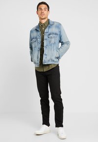 Giacca di jeans leggera blu con due tasche sul petto, abbinata a una camicia verde oliva con bottoni, pantaloni neri slim e sneakers bianche.