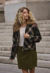 Jauna sieviete ar cirtainiem blondiem matiem, ģērbusies pārbaudītā džaketē ar kažoku, baltā topā un olīvzaļās svārkos, stāv pie akmens kāpnēm.