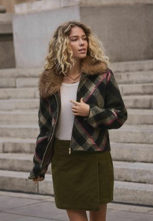 Tânără cu păr blond creț, îmbrăcată cu o jachetă în carouri cu guler de blană, un top alb și o fustă verde olive, stând lângă trepte de piatră.