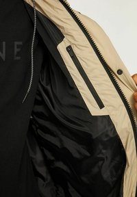 Interno di giacca beige con fodera nera; presenta una tasca con zip e cuciture a vista. Texture morbida e design elegante.