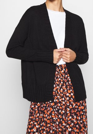 Cárdigan negro de punto, con apertura frontal, puños acanalados y dobladillo. Combinado con una falda floral que presenta patrones naranjas y blancos sobre un fondo oscuro.