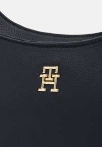 Marineblaue Leder Tasche mit einer genarbten Oberfläche, die ein goldfarbiges, geometrisches Logo-Detail nahe der oberen Kante aufweist. Glatte Kanten und strukturiertes Design.