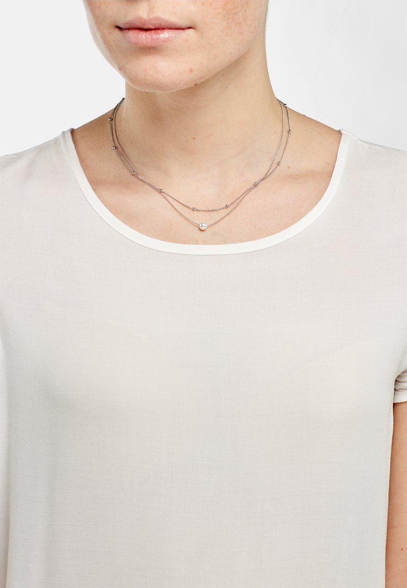 Gelaagde ketting met fijne zilveren kettingen, voorzien van kleine zilveren kralen en een enkele centrale parel, gedragen over een lichte, eenvoudige ronde hals top.