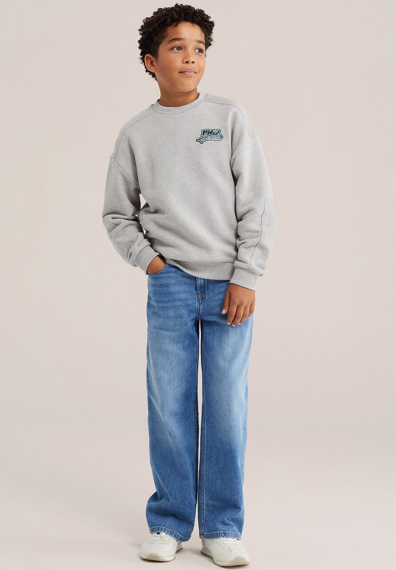 Garçon aux cheveux bouclés portant un sweat gris, un jean bleu à jambes larges et des baskets blanches, debout avec une main dans la poche, regardant sur le côté.