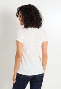 Camiseta de manga corta de color crema con un diseño de puño enrollado. La tela parece suave y ligera, con una textura sutil.