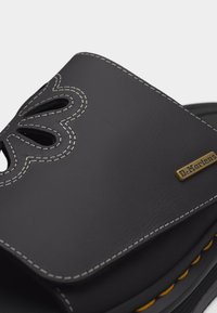 Μαύρο σανδάλι slide Dr. Martens με λευκή ραφή, καρδιοειδείς εξοχές και μικρή μεταλλική ετικέτα με το λογότυπο στο λουρί.