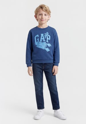 Garçon aux cheveux blonds portant un sweat-shirt bleu avec des motifs de vaisseau spatial et d'astronaute, un jean foncé et des baskets blanches, debout devant un fond blanc.