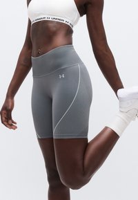 Αθλήτρια με λευκό αθλητικό σουτιέν και γκρι σορτς Under Armour τεντώνει τους τετρακέφαλους, κρατώντας τον δεξί αστράγαλο πίσω με το αριστερό χέρι χαλαρό.