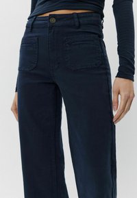 Jūras zila brīva piegriezuma bikses, izgatavotas no kokvilnas kokvilnas maisījuma auduma. Iespējas aizmugurējās kabatas un priekšējā pogas aizdare ar jostu cilpām. Gluda tekstūra.