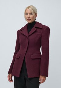 Woman wearing a fitted burgundy blazer with wide lapels, flap pockets, and black trousers, standing against a plain background.