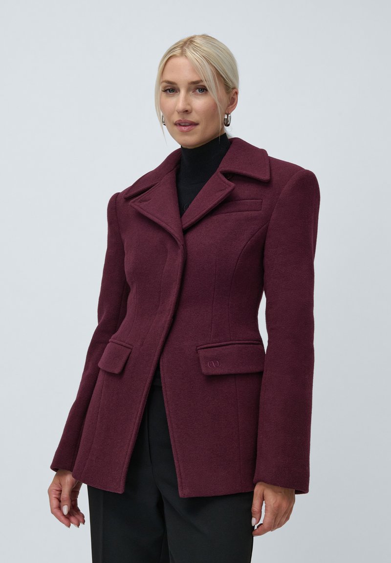 Woman wearing a fitted burgundy blazer with wide lapels, flap pockets, and black trousers, standing against a plain background.