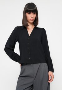 Blusa preta de mangas longas com colarinho, decote em V e oito botões metálicos. Apresenta mangas bufantes e um corte descontraído.