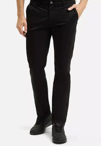 Mężczyzna w slim-fit czarnych spodniach i czarnych trampkach, z jedną ręką w kieszeni, stoi na tle białym.