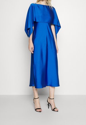 Robe en satin bleu avec une cape structurée, longueur genou, coupe décontractée et légère brillance ; associée à des escarpins noirs à brides.