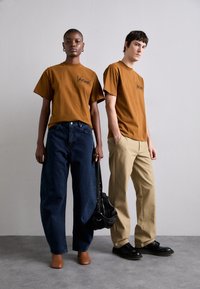 Brązowe koszulki z logo, luźne niebieskie dżinsy, beżowe spodnie i czarne buty; czarna torba trzymana przez modela. Prosty, casualowy styl.