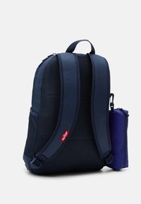 Sac à dos bleu marine avec tissu en mesh, bretelles rembourrées et poche latérale pour une petite trousse violette. Fermeture zippée et poignée robuste sur le dessus.