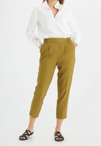 Olijfgroene tapered broek met elastische tailleband, zijzakken en opgerolde manchetten, gecombineerd met een witte lange mouwen blouse met kraag en zwarte sandalen.