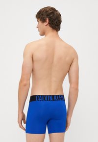 Boxer in microfibra blu con elastico nero sul fondo che presenta la scritta "CALVIN KLEIN" in lettere blu. Il materiale sembra liscio e elasticizzato. Non ci sono loghi visibili sui boxer.