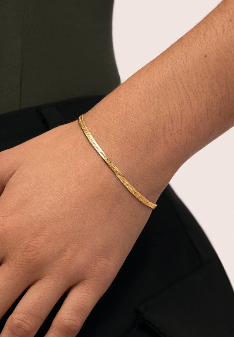 Pulsera tono dorado en una muñeca, con un diseño liso y plano, superficie texturizada y perfil delgado.