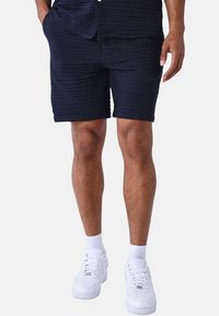 Bermudas de color azul marino con textura y un patrón de acanalado, que presentan dos bolsillos laterales, combinadas con zapatillas blancas y calcetines cortos.