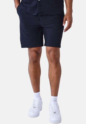 Bermudas de color azul marino con textura y un patrón de acanalado, que presentan dos bolsillos laterales, combinadas con zapatillas blancas y calcetines cortos.