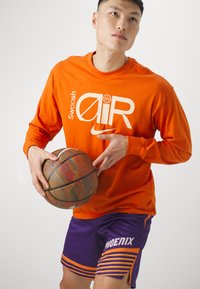 Pomarańczowa koszula z długim rękawem z białymi grafikami, w połączeniu z fioletowymi szortami z pomarańczowymi paskami i napisem "PHOENIX". Trzyma koszykówkę.