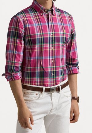 Homme portant une chemise à manches longues à carreaux roses rentrée dans un pantalon blanc avec une ceinture marron, manches retroussées.