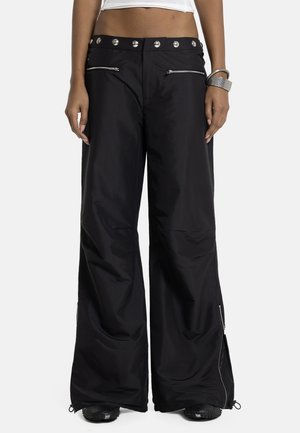 Femeie purtând pantaloni largi negri cu buzunare cu fermoar argintiu și brățări cu cataramă argintie pe încheietura mâinii drepte.