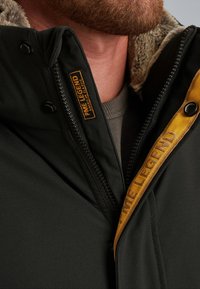 Giacca invernale nera con colletto foderato in pelliccia sintetica, chiusura con zip e etichetta arancione, in tessuto liscio e con rifinitura gialla a contrasto sulla zip.