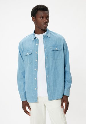 Light blue denim shirt with button closure, two chest pockets, and a pointed collar. Worn over a white t-shirt, casual fit.