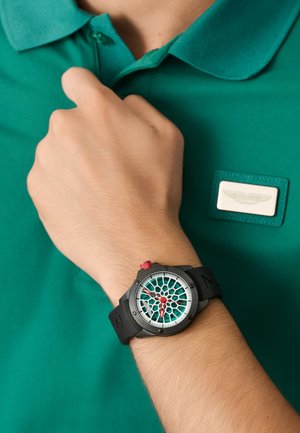 Reloj de pulsera negro con esfera de patrón verde azulado y corona roja. Lleva puesto en una mano contra una camiseta polo verde con un parche de logo blanco.