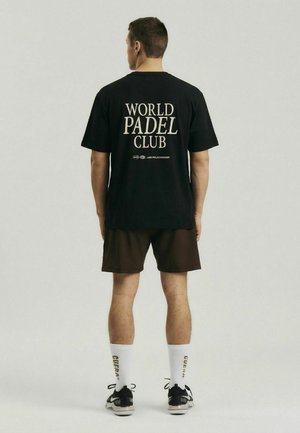 Mand stående med ryggen til iført sort "World Padel Club" t-shirt, brune shorts, hvide sokker med tekst og sorte sneakers på ensfarvet baggrund.