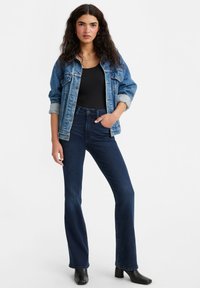Denim jacket in blauw met een ontspannen pasvorm, opgerolde mouwen, zwart topje, donkerblauwe flared jeans en zwarte enkellaarzen. Eenvoudig ontwerp met minimale hardware.